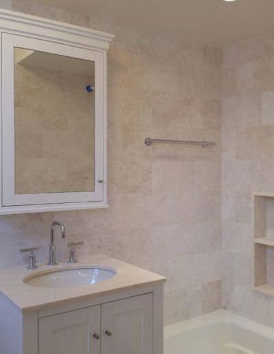 A modern bathroom with a beige tile finish, featuring a sink vanity, mirror cabinet, bathtub with a grab bar, and shelving nook.