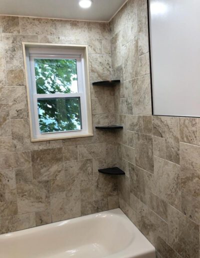 A modern bathroom with beige tiled walls, a white bathtub, and built-in corner shelves with a window overlooking greenery.