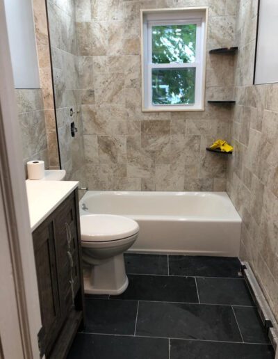 A small bathroom with beige tiled walls, a white bathtub to the right, a toilet, and a dark-tiled floor with a window providing natural light.