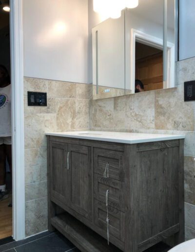 A bathroom vanity with a marble countertop and wooden cabinetry, featuring a mirror above and a partial reflection of a person in the doorway.