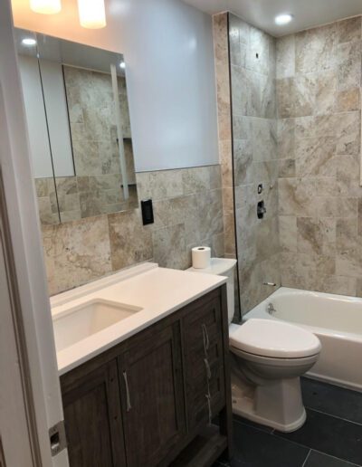 Modern bathroom interior with stone tiles, featuring a vanity cabinet, mirror, toilet, and bathtub.