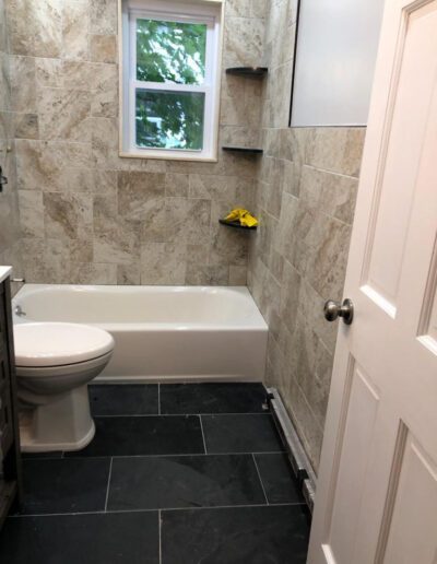 A bathroom with beige tiled walls, black floor tiles, a white bathtub, toilet, and a small window with a view of greenery outside.