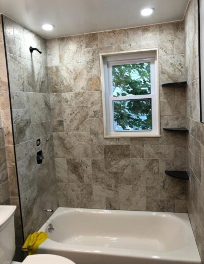 Modern bathroom with beige tiled walls, a white bathtub, and a window with a view of trees.