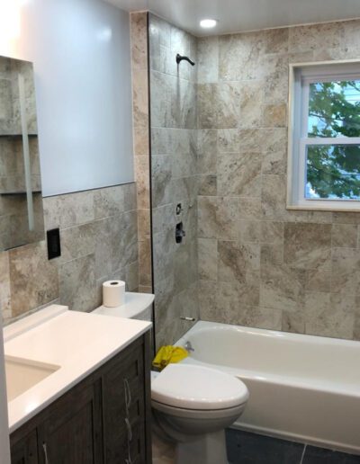 A modern bathroom with beige tiles, a shower tub combo with glass doors, a white sink, a mirrored cabinet, and a window.