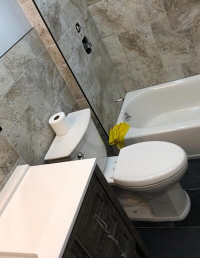 A modern bathroom interior featuring a toilet with a roll of paper on top, a vanity cabinet, and a bathtub with a yellow cloth hanging on the side.