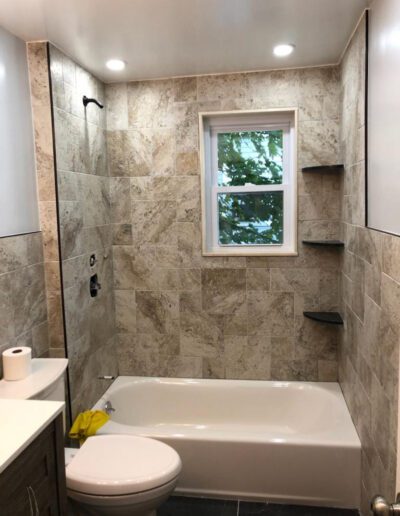 Modern bathroom with beige tiles, a bathtub, and a window facing greenery.