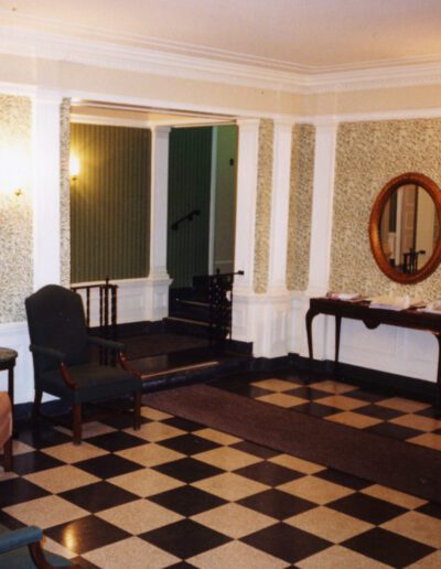 An interior view of a vintage room with checkered flooring, patterned wallpaper, antique furniture, and wall sconces.