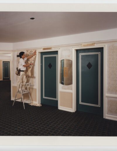 Two workers installing wallpaper in a hallway with two elevator doors.