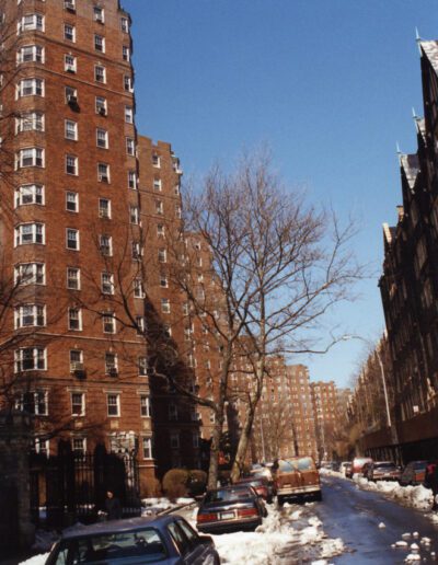 A tree-lined street with parked cars and snow on the sides in front of a traditional brick apartment building on a clear day.