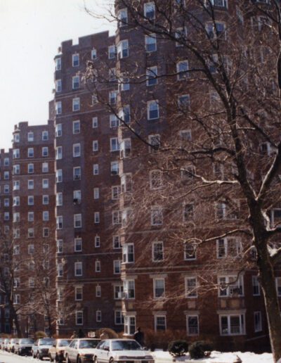 Brick apartment buildings line a snow-covered street with parked cars.