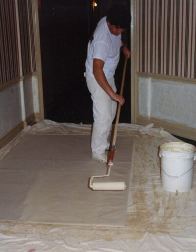 A person applying primer or paint on a large canvas on the floor, with painting supplies nearby.