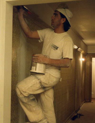 Man applying wallpaper in a corridor while another person works in the background.