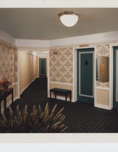 An empty hotel corridor with patterned wallpaper, doors with diamond-shaped panels, and benches with a potted plant.