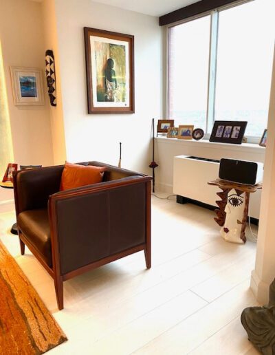 A cozy home office with a leather armchair, desk with computer, artwork, and a window for natural light.