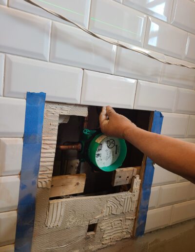 A person's hand adjusting a green valve through an opened tile wall.