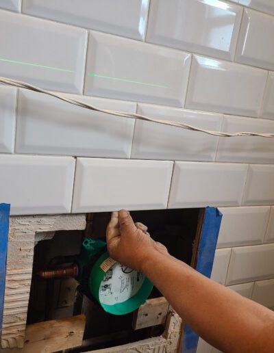 Person installing white subway tiles using a laser level guide around a kitchen backsplash area with an exposed section for plumbing.