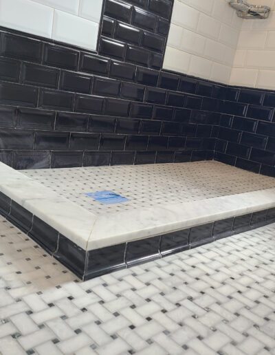 A modern bathroom corner with black subway tiled walls and a white mosaic tiled floor, featuring a built-in shower area without fixtures.