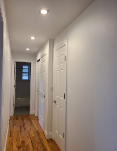 A well-lit hallway featuring wooden flooring, white walls, and closed white doors leading to different rooms.