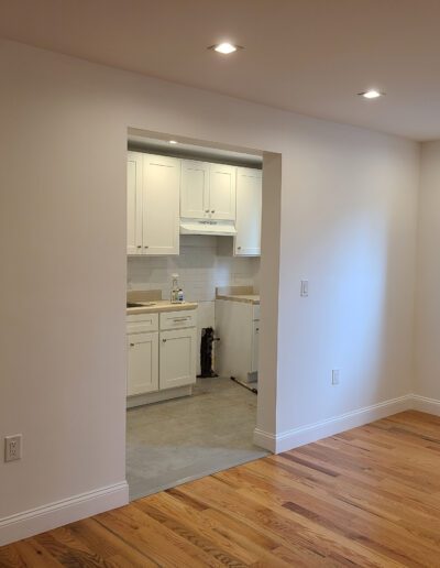 A clean, empty room with hardwood floors, white walls, and a view into a small white kitchen.