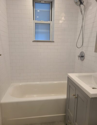 A modern bathroom with white subway tiles, featuring a bathtub with an overhead shower, a small window, and a gray vanity sink.