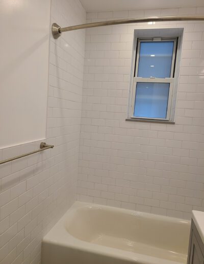 A clean, white-tiled bathroom with a bathtub and a small window letting in daylight.