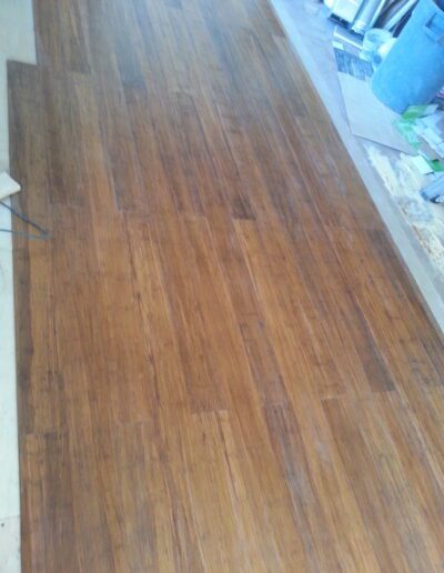 A wooden floor with a warm brown finish, showing signs of use, with some clutter visible in the background.