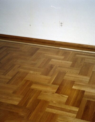 Corner of a room with herringbone parquet flooring and baseboard with a visible date stamp on the photo.