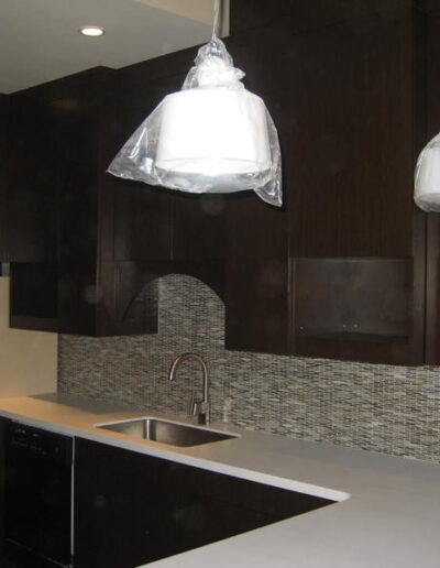 Modern kitchen with dark wood cabinets and mosaic backsplash, featuring a sink and two pendant lights covered in protective plastic.