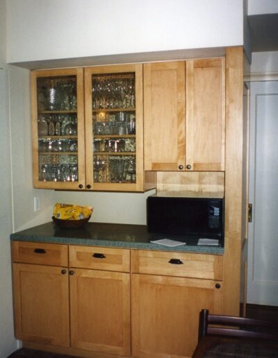 A traditional kitchen corner featuring wooden cabinetry with glass door fronts, a countertop with a microwave and a bowl of fruit, and patterned flooring.