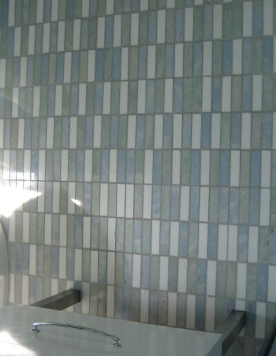 Interior corner showing a patterned tile wall with sunlight casting a glare, next to a metal table with a glass partition.