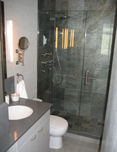 Modern bathroom with a glass shower enclosure, gray tiles, and a white toilet beside a vanity with a mirror.