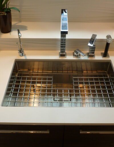 A modern kitchen sink with a stainless steel faucet and grate, set in a white countertop with wood cabinets below.