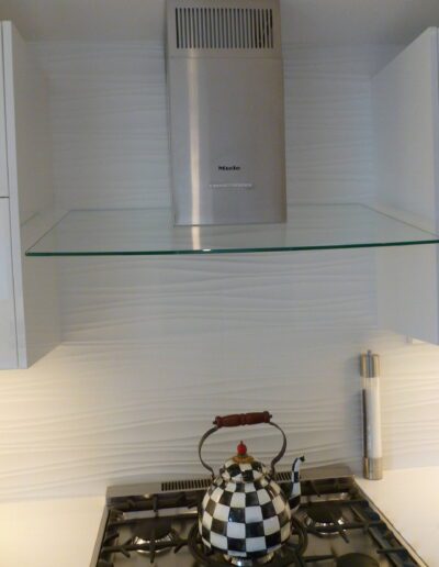 A modern kitchen stovetop with a checkered kettle below a stainless steel range hood and a floating glass shelf.