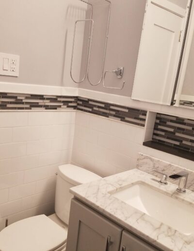 A modern bathroom with a marble countertop sink, white toilet, and tiled walls featuring a decorative mosaic accent band.