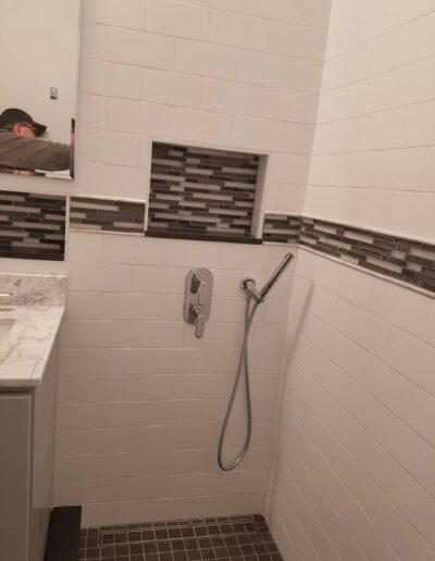 A modern bathroom corner with white subway tiles, featuring a built-in shelf with mosaic accents, a wall-mounted shower faucet, and a hand-held shower head.