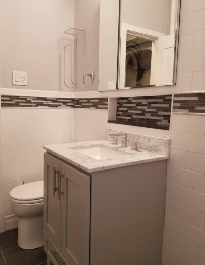 A modern bathroom interior featuring a white toilet, a gray vanity with a marble countertop, and a reflection of a shower in the mirror.