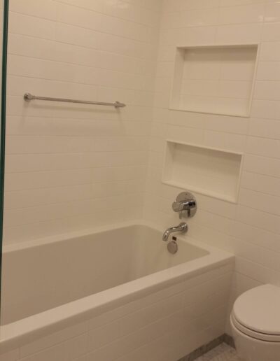 A clean, white-tiled bathroom with a bathtub, built-in shelving, and a toilet.