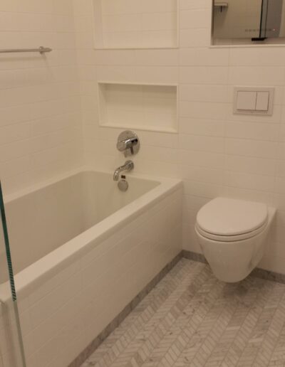 A modern bathroom with a white bathtub, toilet, and herringbone tile flooring.