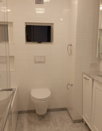 Modern bathroom interior featuring white tiles, a wall-mounted toilet, and a vanity with a reflection of a window at dusk.