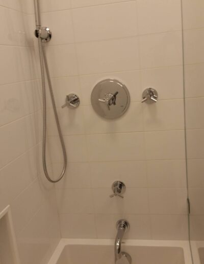 A modern shower with white tiles, featuring a handheld showerhead and multiple control valves.