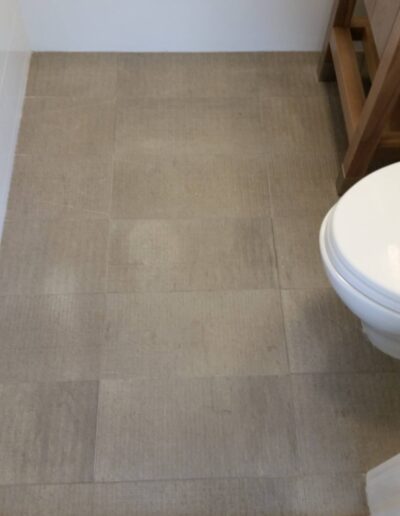 A bathroom floor with beige tiles extending to a wooden threshold, featuring a partial view of a toilet and a wooden vanity to the left.