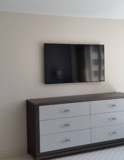 A modern bedroom interior with a mounted television above a gray dresser, adjacent to an open door leading to another room.