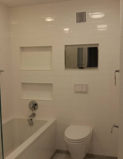 A modern bathroom with white tiled walls, a bathtub with a shower, a toilet, and built-in wall shelves.