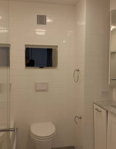 Modern bathroom interior with white tiles, featuring a toilet, sink, and a mirror reflecting a television screen.